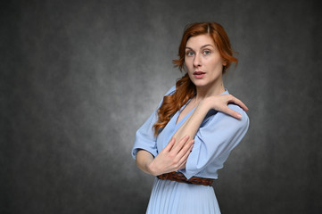 Portrait of a young caucasian woman with long red hair in a blue dress. The model poses with different emotions on a gray background in the studio.