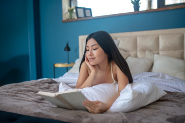 Young brunette lying on bed in night dress, reading book, resting her head on one hand