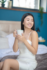 Young brunette in night dress sitting on bed, holding white cup, drinking, smiling