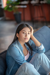Fototapeta premium Upset dark-haired female sitting with folded knees on sofa, thinking, resting her head on one hand