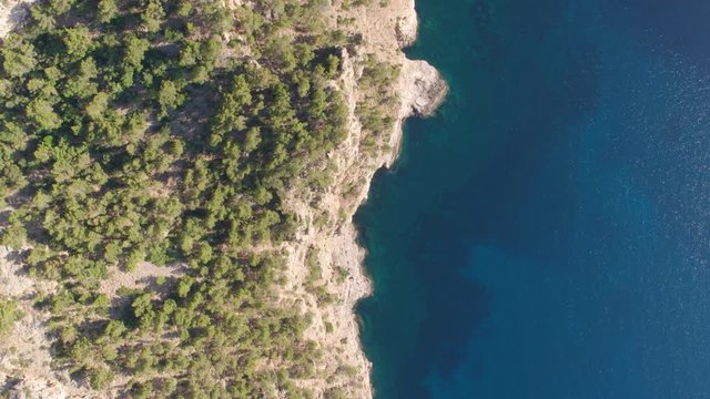 AERIAL DRONE TOP DOWN ZENITHAL SMOOTH SHOT OF MEDITERRANEAN CLIFF SEA SIDE WITH BLUE TURQUOISE WATER AND GREEN PINE FOREST