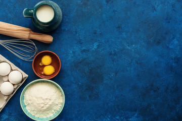 Obraz na płótnie Canvas Baking background. Flour, egg, milk, rolling pin, whisk on a blue painted background. Top view, flat lay, copy space.