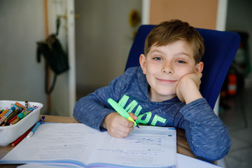 Hard-working happy school kid boy making homework during quarantine time from corona pandemic disease. Healthy child writing with pen, staying at home. Homeschooling concept