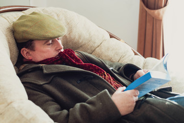 Middle-aged man bored by reading a book at cold home. He falls asleep in his chair