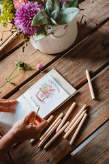 female artist draws a composition of wild flowers in a pot in a rustic style, a sketchbook on a table with pencils.