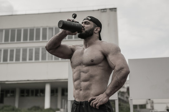 young strong athlete male drinking water from black bottle during outdoor fitness workout on university building backgroung