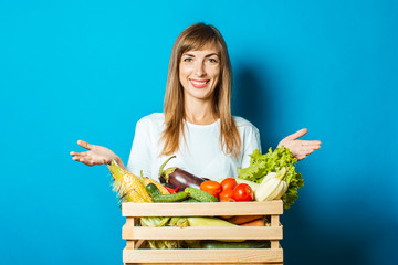 Young woman smiles and holds a box with fresh vegetables on a blue background. Good harvest concept, natural product