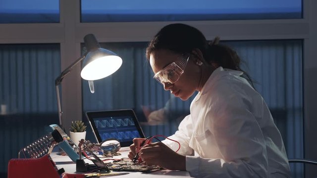Side view of female electronics engineer working with multimeter tester and other electronic devices in laboratory