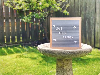 Love your garden. Gardening concept with felt message board on a bird bath in the middle of a suburban garden with dwarf apple tree in the background and copy space. 