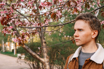A young man with blond hair in a brown jacket and a backpack walks in a blooming garden, spring blooming pink cherry trees, close-up portrait