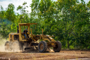 Motor grader clearing and leveling construction site surface with forest in the background. Grader industrial machine on road construction work.