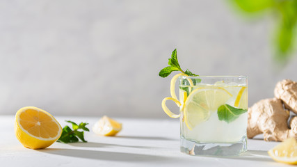 Refreshing lemonade with lemon, mint, ginger and ice in glass and bottle on white concrete background. Summer refreshing drink. Cold detox water with lemon. Copy space. Menu