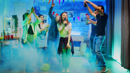 Cheerful young woman singing for her friends at the party in a room with neon lights and smoke.