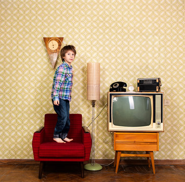 Vintage Art Portrait Of Liitle Boy Jumping On Armchair In Room With Interior From 70s 20th Century, Retro Stylization, Image Toned. Child At Home With Old Tv Set