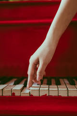 a close-up of a side view with female hand with pink manicure playing the piano. An old red piano with black and white keys. A palm above the keyboard. Graceful fingers of composer or musician
