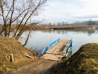 Fototapeta na wymiar Ferry for transporting goods and people across the river moored along the river bank with a lowered ramp ready for loading. Raft made of metal with metal fences and wooden floor.
