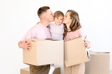 Fototapeta na wymiar Happy family life. Young parents and their little son are standing holding cardboard boxes. They moved to their new own home and kisses their beloved son