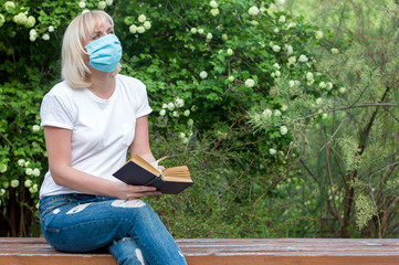 Girl woman in a park on a bench in a medical mask reads a book, walk in the period of quarantine and social isolation, prevention of allergies in the period of seasonal flowering, copy space for text