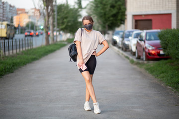 beautiful girl in a mask with a phone and a backpack posing standing on the street and looking at the camera