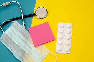 Stethoscope, tablets and pink sticky note for text above face mask on colorful background.