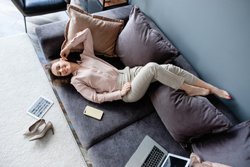 Top view happy healthy young smiling woman enjoying weekend and forced quarantine while lying on cozy sofa at home. Concept of pleasure social media communication and online shopping