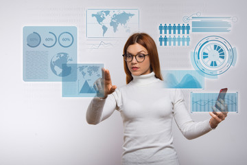 Woman holding smartphone and touching virtual global infographics. Focused young woman in eyeglasses using mobile phone. Technology and advertisement concept