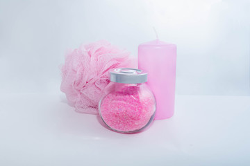A transparent jar with bath salt with a metal lid, a sponge for bath and a candle stand nearby on a white background. All objects have a light pink color. The concept of items for shower, personal