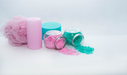 Obraz na płótnie Canvas Two cans of bath salt of light pink and turquoise color, a turquoise jar of cream, a light pink candle and a sponge for a shower lie next to a white background. Body cosmetics and spa items.