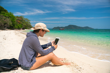 Beautiful Hiking girl with a backpack sittit on a sandy tropical beach and uses a tablet and internet for work.