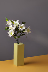 Minimalistic shot of a yellow rectangular vase with a bunch of white lilies in it. The low vase is located on the orange table against the gray wall.  