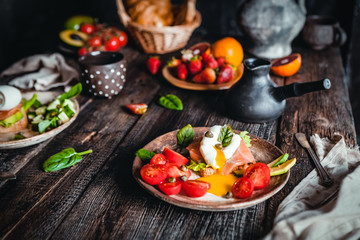 breakfast bowls with poached eggs, vegetables and salmon, oranges and fresh strawberry in plate with mugs of coffee on brown wooden table