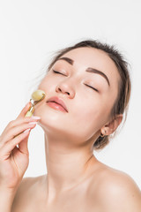 Obraz na płótnie Canvas Beautiful model woman with healthy fresh clean skin enjoying a massage with a jade face roller to improve circulation, relax the muscles and tone the skin, isolated on gray background with copy space