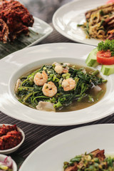 indonesian stir fried water spinach or kangkung  with shrimp on top and blurred food in the background