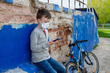 A young European boy is happy to communicate with friends online from a smartphone. A child is sitting on a blue wall in a medical mask from the virus, next to a Bicycle.