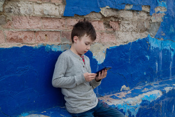 A teenage American boy carefully reads an article on the Internet from a smartphone. The child is sitting on a blue wall.