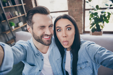 Close up photo of carefree playful positive two people man woman travel trip make selfie show tongue out wink blink sit couch in house indoors