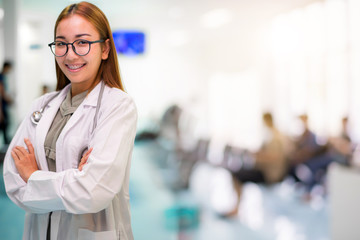 Attractive smiling Asian doctors wearing stethoscope with blur patient crowd background in hospital, during coronavirus or covid-19 crisis, medical concept.
