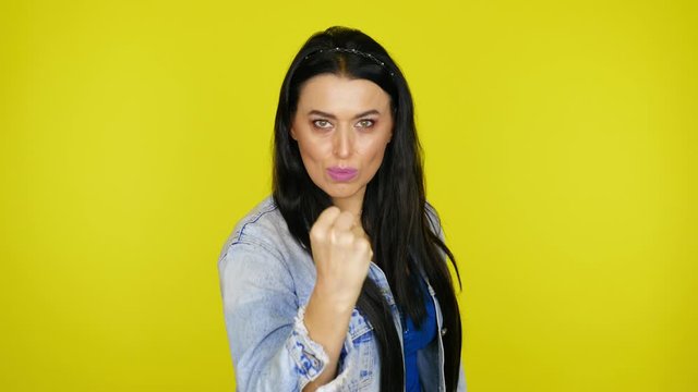 Woman with black hair in a denim jacket shows a fist at the camera on a yellow background with copy space. Place for text or product. 4K footage