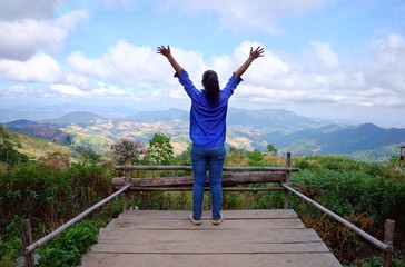 The back view of a woman with blue shirt and jeans looking over a lush green mountains and valley at the edge of a wooden ledge with her arms up, feeling happy and relax after a long journey.