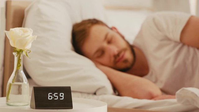 Selective focus of clock and rose on bedside table and man sleeping at morning
