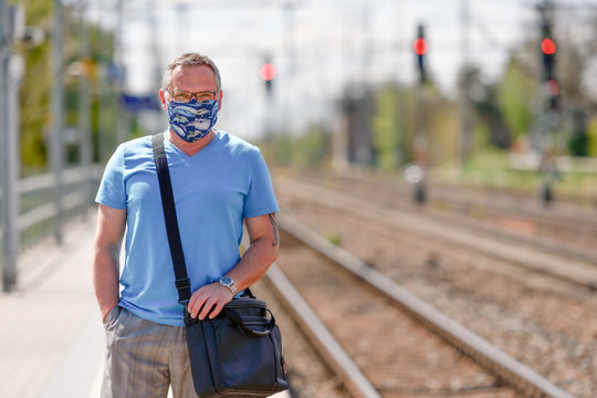 Mann mit Mund-Nasenschutz wegen der Corona Krise wartet auf dem Bahnhof auf den Zug-Gleise im HIntergrund, in die Kamera schauened