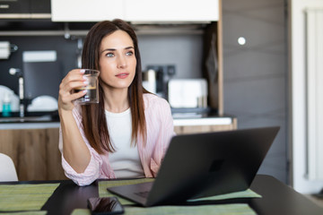 Young woman using a laptop while drinking a cup of a coffee in her kitchen