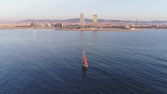 AERIAL DRONE SMOOTH ORBIT OF SAILING BOAT DURING SUNRISE WITH BARCELONA IN THE BACKGROUND AND CALM BLUE SEA 