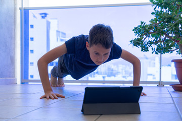 Kid with tablet computer doing push-ups sport exercises on balcony. Sport, healhty lifestyle, training, active leisure at home