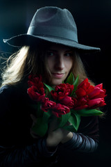 The girl in the purple dress and wide-brimmed black hat on a dark background in the Studio with a red Tulip flower in the hand closes with a Bud eye