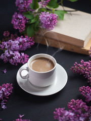 Obraz na płótnie Canvas a white Cup of fragrant coffee on a dark table with lilac bush and a book. romantic home atmosphere