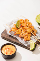 prawns on skewers with lime and sauce on parchment paper on wooden board on white background