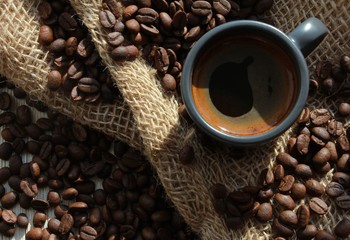 Grey cup of fresh coffee with foam, coffee beans on rough burlap jute sack. Closeup, top view. Agriculture, traditional coffee shop concept