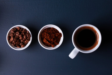 Espresso white cup, whole roasted beans and ground coffee powder on dark blue table. Stages of coffee preparation, line of cups. Top view, arrangement. Coffee shop, morning concept