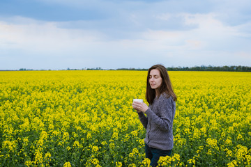 Fototapeta premium The girl stands in the middle of a flowering rapeseed field and holds a cup of coffee in cloudy weather 